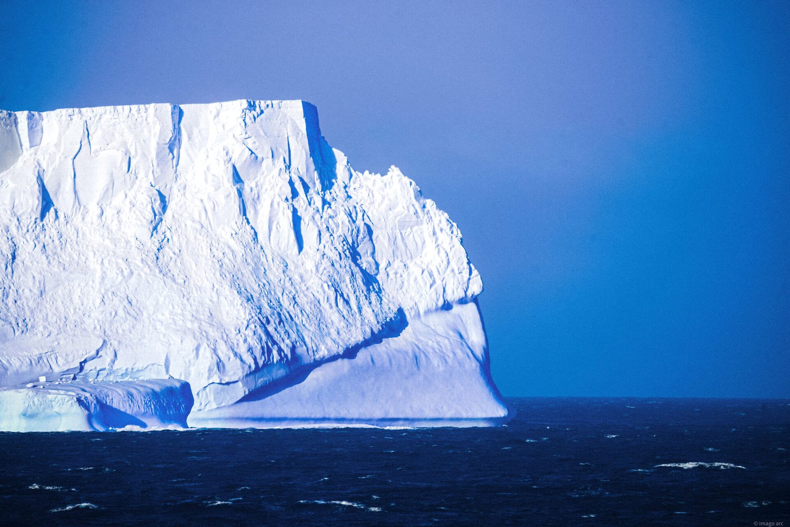 Massive iceberg wall lit in blue and white against a clear blue sky, Antarctica