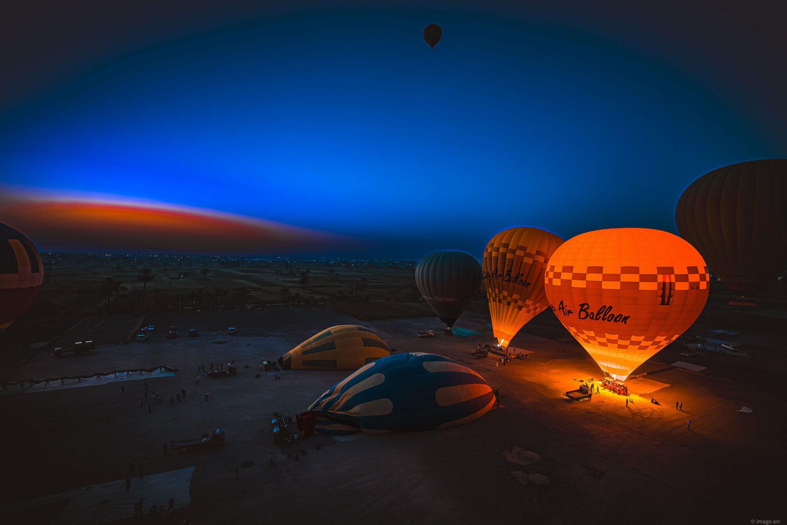 Hot air balloons launching at pre-dawn over Luxor, Egypt, with glowing burners against a deep blue sky